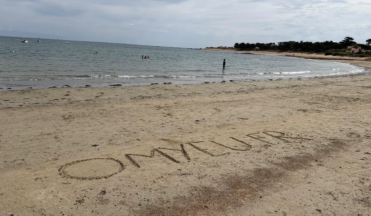 omyeu.fr écrit en grandes lettres dans le sable d'une plage, la mer et des bateaux derrière